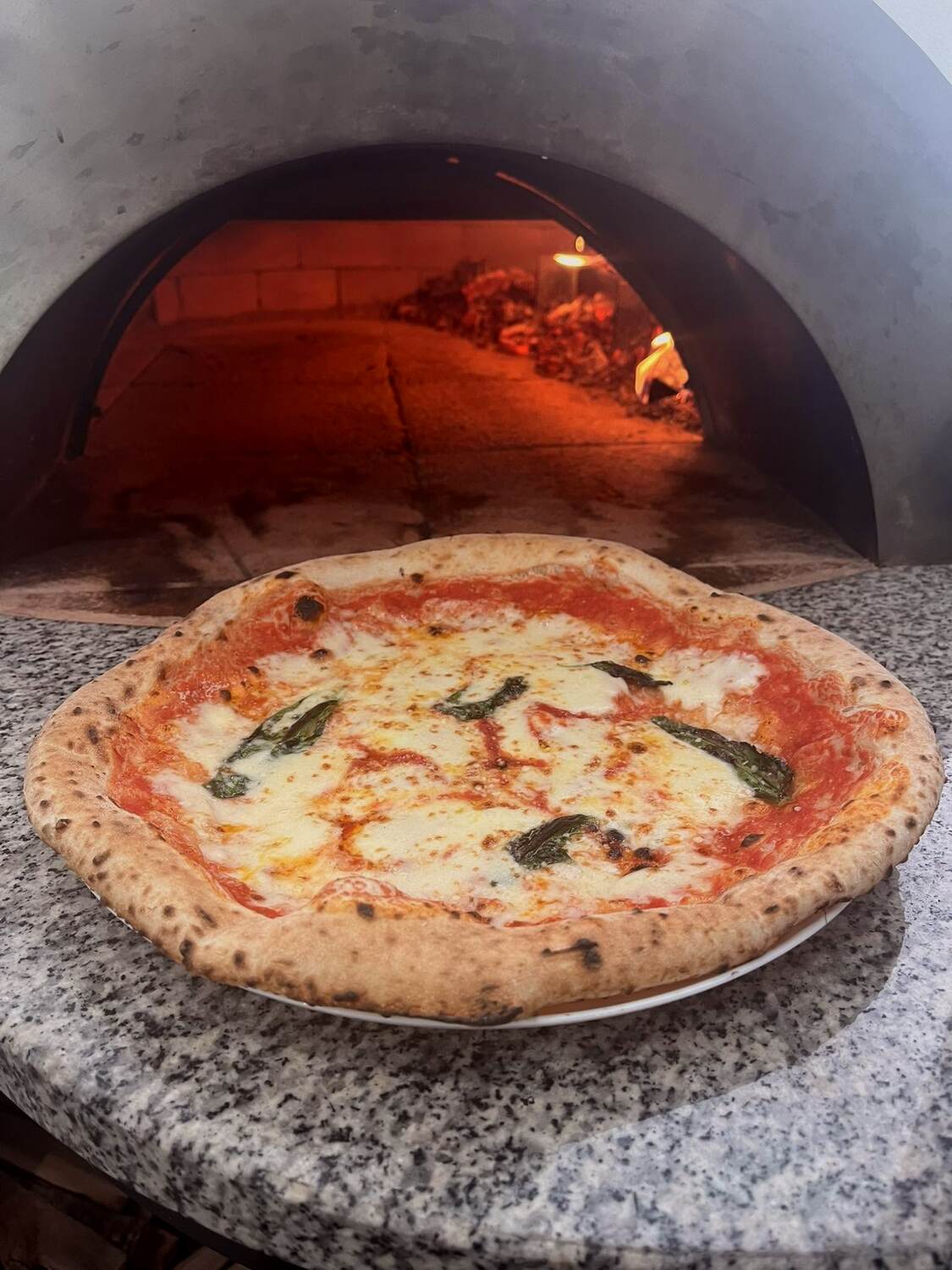 Egész margherita pizza tányéron, a háttérben a kemence parázsával