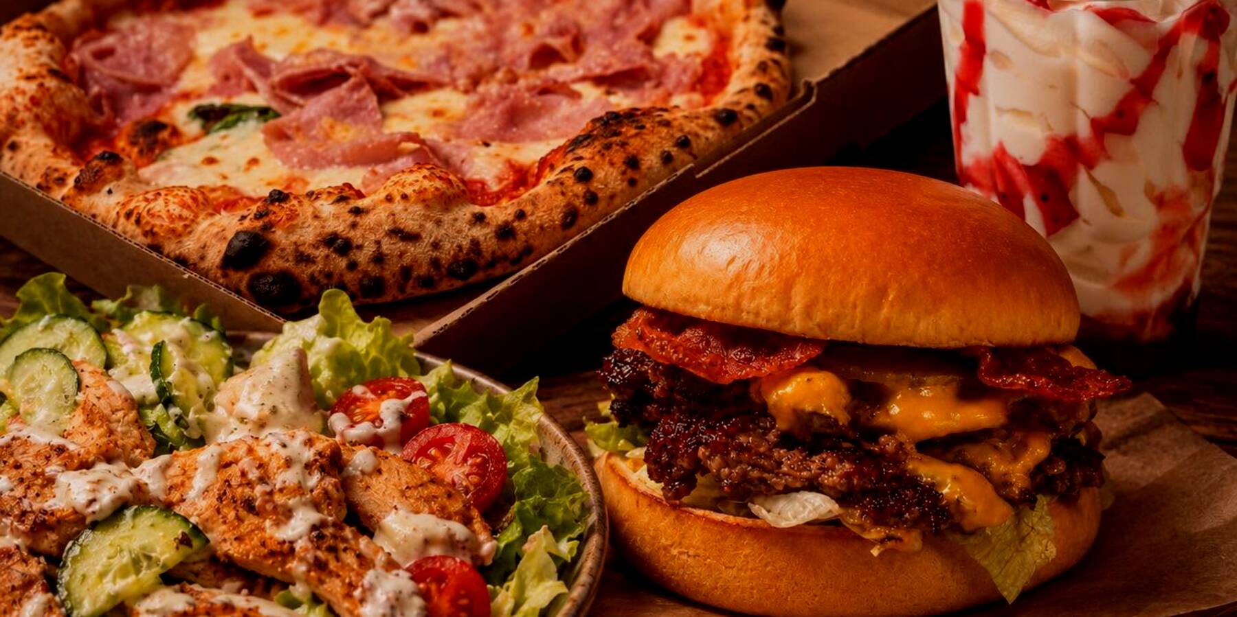 Pizza dobozban, burger deszkán és saláta egy asztalon